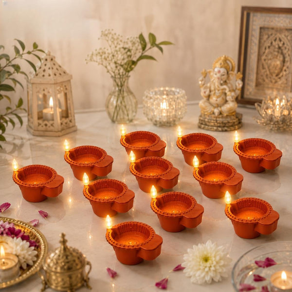 A styled arrangement of orange clay diya candles lit with small flames, placed on a light marble surface with decorative props including a lantern, flowers, a brass lamp, and a Ganesha statue in the background.